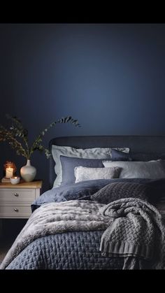 a bedroom with dark blue walls and grey bedding in the foreground is a white table with two vases on it