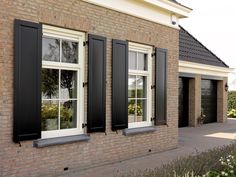 two windows with black shutters on the side of a brick building next to flowers