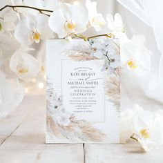 a white wedding card with flowers and lights in the background on a wooden table next to a bouquet of orchids