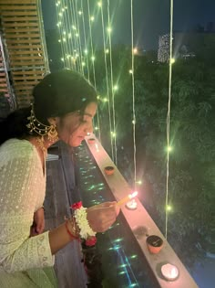 a woman lighting candles on a balcony at night with lights strung from the ceiling and trees in the background