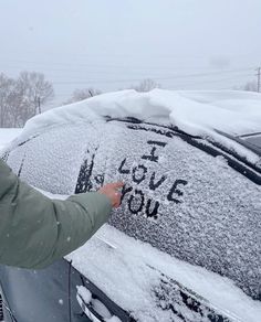 a person writing on the windshield of a car covered in snow, with trees and buildings in the background