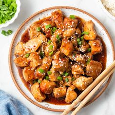 a white plate topped with meat covered in sesame seeds and green onions next to chopsticks