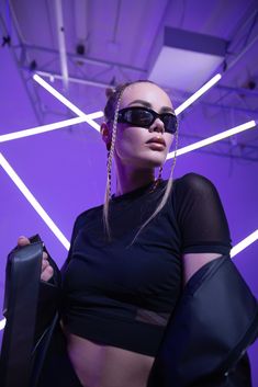 a woman wearing sunglasses and holding a black bag in front of a purple wall with neon lights