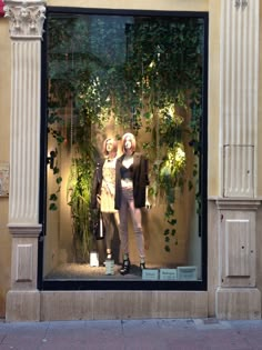 two mannequins are standing in front of a store window with greenery