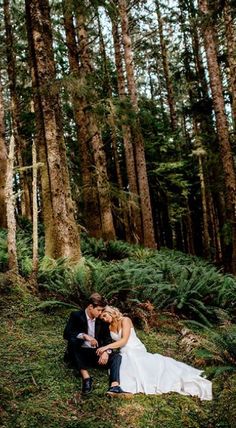 a bride and groom sitting on the ground in front of some tall trees at their wedding