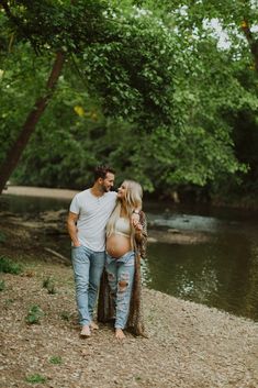 a pregnant couple standing next to each other in front of a river with trees around them