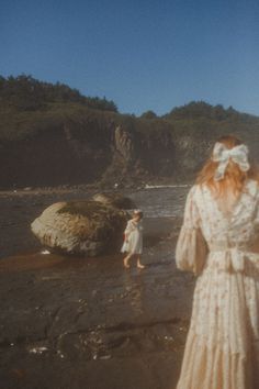 a woman in a white dress is walking on the beach with another person behind her