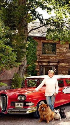 a woman standing next to two dogs in front of a red car