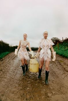 two women in short skirts and boots are walking down a dirt road carrying a yellow container