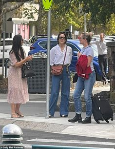 three women are standing on the sidewalk near a street sign and suitcases, talking to each other