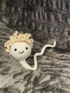 a crocheted doll with blonde hair laying on a gray bed sheet and looking at the camera