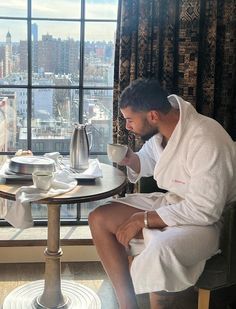 a man sitting at a table in front of a window drinking from a coffee cup