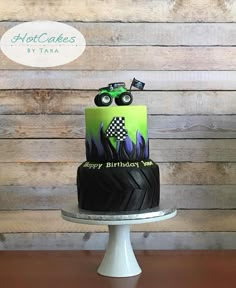 a green and black birthday cake with monster trucks on it's side, sitting on top of a white pedestal