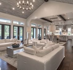 a living room filled with white couches and chairs next to a kitchen area covered in windows