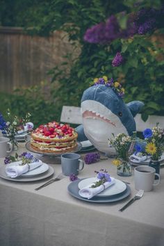 a table topped with plates and cups next to a stuffed animal shark on top of a cake