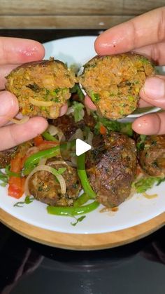 two hands holding food over a white plate with vegetables and meatballs on the side