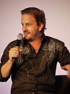 a man holding a microphone in his right hand and wearing a black shirt with an intricate design on it