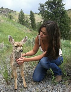 a woman petting a baby deer on the side of a hill with trees in the background