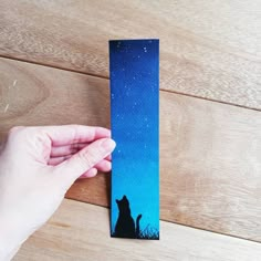 a hand is holding up a bookmark with a cat on it and the sky in the background