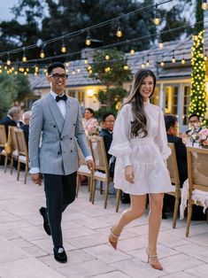 a man and woman walking down the street in front of some tables with lights on them