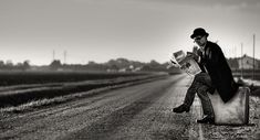 a man sitting on top of a suitcase reading a newspaper in the middle of an empty road