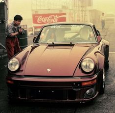 a man standing next to a red sports car in the rain with coca cola cans behind him