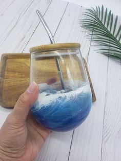 a hand is holding a glass with blue liquid in it on a wooden tray next to palm leaves