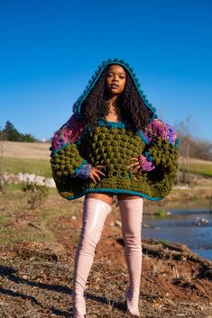 a woman wearing a green crocheted sweater and pink pants stands in front of a pond