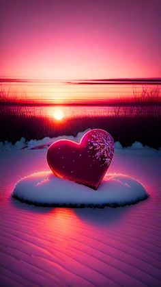 a red heart sitting on top of snow covered ground next to the ocean at sunset