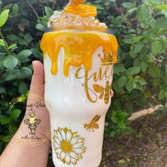 a hand holding a yellow and white glass with honey on it's side, in front of some bushes