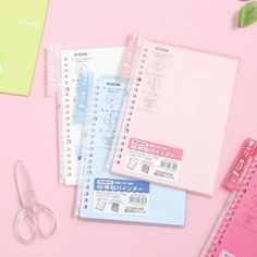 various stationery items laid out on a pink surface with scissors, notebooks and pens