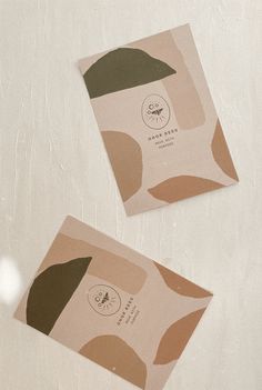 two square business cards on top of a white surface with brown and green shapes in the middle