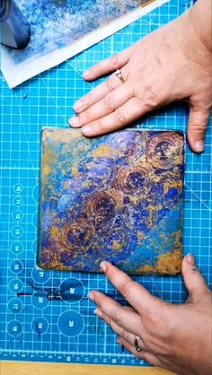 two hands are working on a piece of art that is being made with blue and gold paint