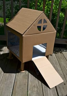 a cardboard dog house with the door open on a wooden deck in front of a fence