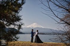 a couple standing next to each other in front of a lake with a mountain in the background