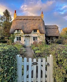 a thatched roof house with a white picket fence in the foreground and green bushes on either side