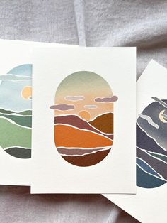 three cards with mountains, clouds and the sun in them are on a bed sheet