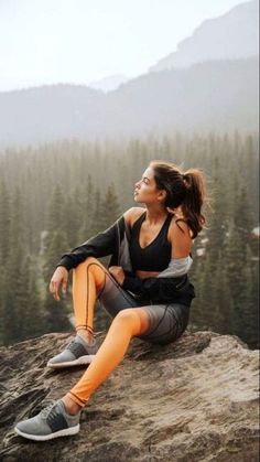 a woman sitting on top of a large rock next to a tree filled mountain range