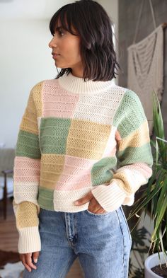 a woman standing in front of a plant wearing a multicolored sweater and jeans