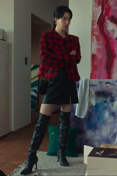 a woman standing in front of a painting wearing black boots and a red sweater with her arms crossed