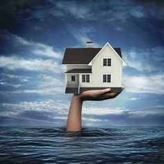 a hand holding a house in the middle of water with blue sky and clouds behind it