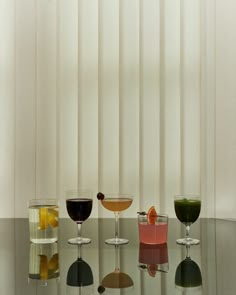 four glasses of different types of drinks on a glass table with white vertical blinds in the background