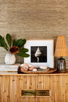 a wicker sideboard with pictures and shells on it, next to a lamp