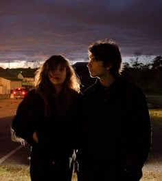 two people standing next to each other on the side of a road at night time