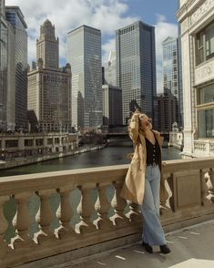 a woman standing on the side of a bridge talking on a cell phone in front of a cityscape