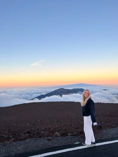 a woman standing on the side of a road with clouds in the sky behind her