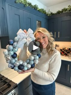 a woman standing in a kitchen holding a wreath with ornaments on it and smiling at the camera