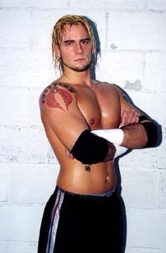 a shirtless man with his arms crossed and tattoos on, leaning against a white brick wall