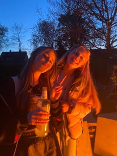 two young women standing next to each other holding drinks in front of a fire pit