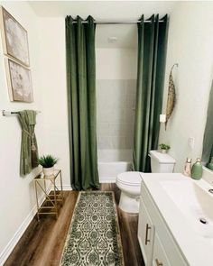 a white bathroom with green curtains and rugs on the wooden floor next to the toilet